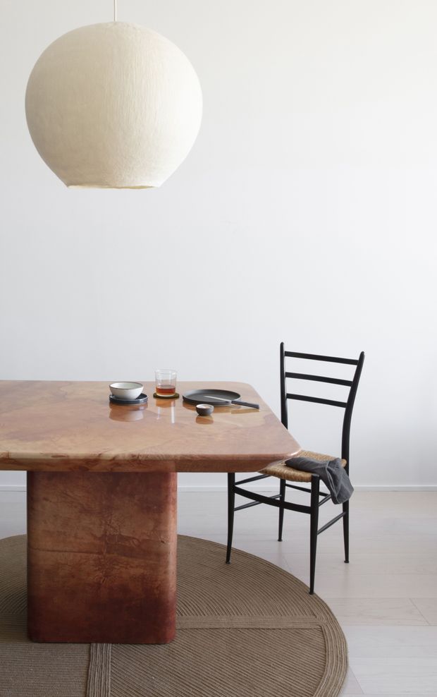 A minimalist dining area with a sculptural warm-toned timber dining table, black ladder-back chairs, and a large textured spherical pendant light. The room has white walls, pale timber flooring, and a neutral woven rug, creating a restrained contemporary setting.