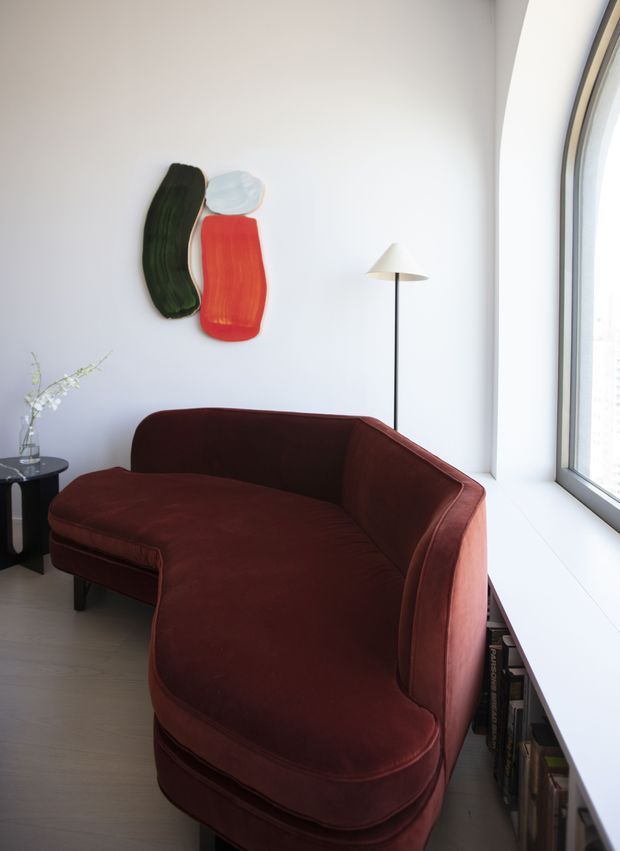 A bright, compact living room corner with a deep burgundy curved sectional sofa positioned beside a large floor-to-ceiling window. A slim black floor lamp, small round side table with a vase, colorful abstract wall art, and a built-in low window ledge with books complete the space. The room has white walls and pale timber flooring with a refined contemporary feel.