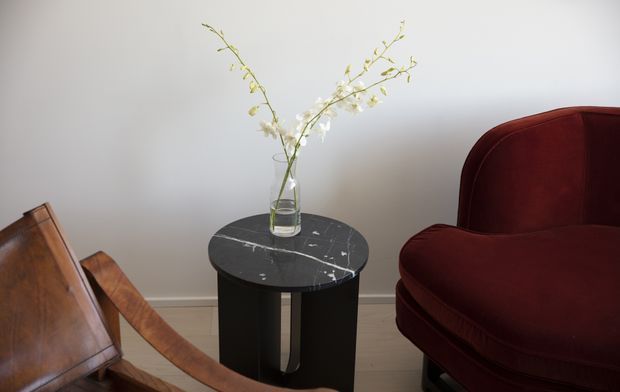 A refined interior vignette featuring a round black marble side table with a glass vase of delicate branches, positioned between a deep burgundy upholstered armchair and a warm-toned wooden chair. The room has pale neutral walls and light timber flooring, creating a quiet, elegant atmosphere.
