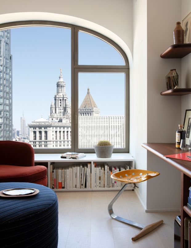 A bright, compact urban home office or reading nook with a dramatic arched window overlooking a city skyline and historic civic buildings. The room includes built-in low book storage beneath the window, a deep red upholstered lounge chair, a dark blue ottoman, a small orange sculptural chair, and dark wood wall shelving and desk joinery. The white walls and pale timber floor create a clean, refined interior with strong architectural window detailing.