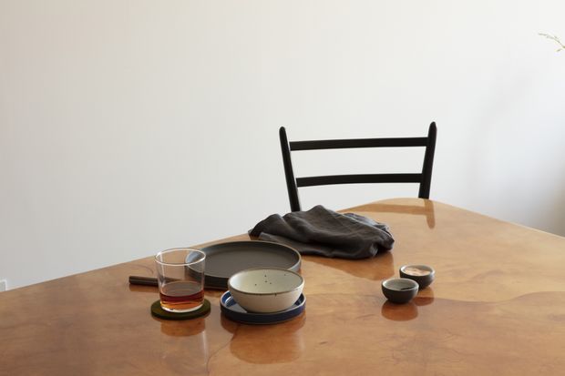 A minimalist dining area centered on a warm timber dining table, with a black ladder-back chair, ceramic cups and plates, a glass, and a folded dark textile. The scene has soft natural light and a restrained, uncluttered composition.