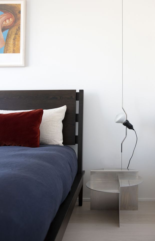 A close-up view of a refined modern bedroom featuring a dark timber bed frame and headboard, navy bedding, a deep red velvet cushion, and a light textured cushion. A sculptural metallic bedside table supports a minimalist wall-mounted pendant light, while white walls, pale flooring, and framed artwork create a calm, contemporary setting.