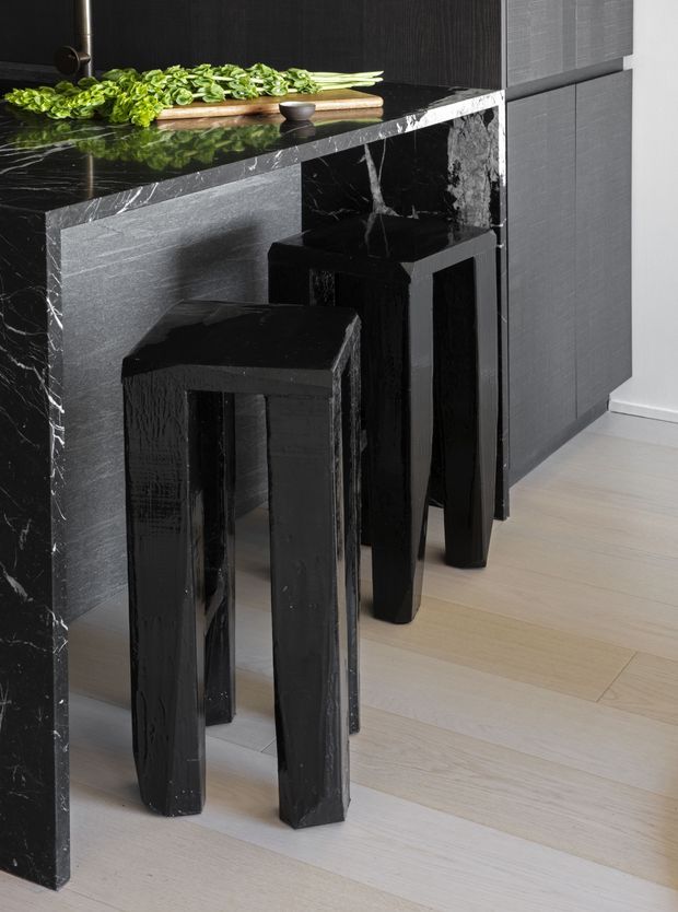 Close-up of a refined dark kitchen or bar counter with a dramatic black marble waterfall edge and matching dark cabinetry. Two tall black bar stools sit beneath the counter, while pale timber flooring provides contrast. Fresh green produce and a timber cutting board rest on the polished benchtop.