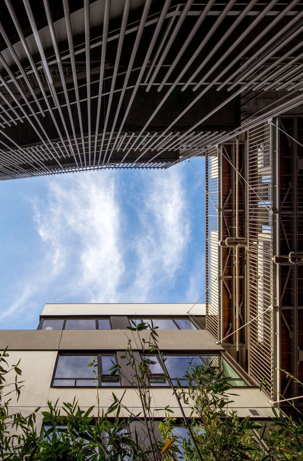 Upward-looking architectural photograph of a contemporary multi-storey building enclosing an open courtyard or narrow urban void. The facade combines pale rendered walls, dark-framed glazing, extensive horizontal metal louvres and projecting linear screens, with exposed service pipes and structural elements visible on the right. Vegetation rises from the lower foreground, while blue sky occupies the central opening. The image strongly emphasizes layered shading devices, facade articulation and a vertical courtyard-like space.