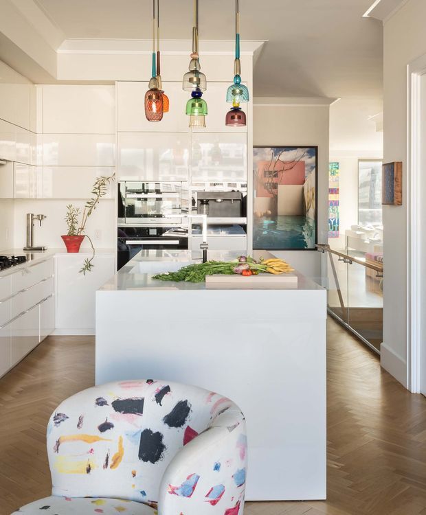 Bright contemporary kitchen with a large white island, glossy white cabinetry, integrated appliances, timber herringbone flooring, and colourful pendant lights. An abstract artwork and mirrored surfaces add visual interest, while a patterned accent chair occupies the foreground.