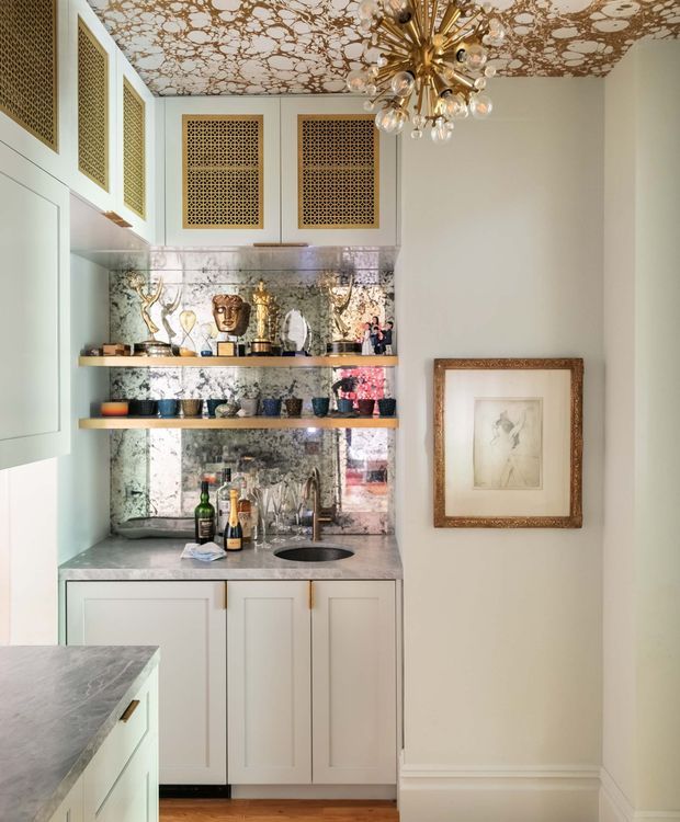 A compact, highly styled home bar or drinks nook with pale framed cabinetry, brass lattice cabinet inserts, open display shelves, a mirrored metallic backsplash, stone benchtop, integrated sink, and a sculptural brass-and-glass starburst chandelier. The adjacent wall is decorated with a framed artwork, while timber flooring and refined trim details add warmth and a traditional luxe character.