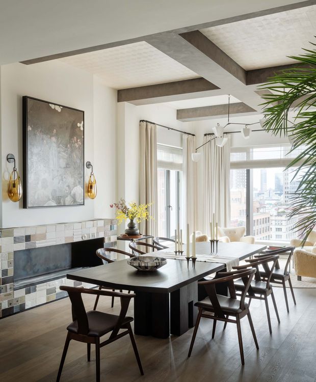 Bright, refined dining room with a large dark timber dining table and matching mid-century-style chairs arranged in front of a tiled fireplace. Floor-to-ceiling windows with sheer curtains overlook a dense urban skyline. The room features exposed concrete ceiling beams, a patterned tiled fireplace surround, pendant lighting, framed artwork, and indoor palms.