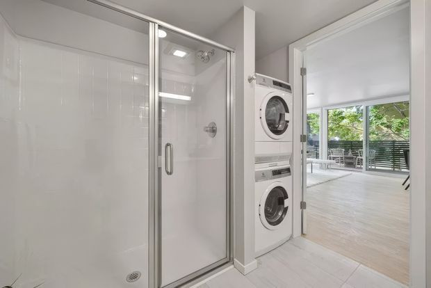 Bright, compact laundry area with a glass-enclosed shower on the left and stacked white washer and dryer beside it. A doorway opens to a light-filled living or outdoor-adjacent space with large glazing and a balcony or terrace beyond. The room has white tiled walls and floor, white cabinetry or appliances, and a clean contemporary appearance.