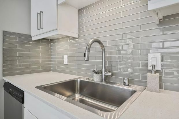 Close-up view of a bright, contemporary kitchen or utility-room sink area with a large stainless-steel double-bowl sink, curved chrome faucet, pale engineered-stone benchtop, glossy grey subway-tile splashback, white cabinetry, and a visible dishwasher.