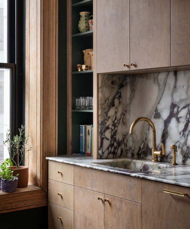 A close-up view of a refined kitchen or kitchenette with full-height light timber cabinetry, integrated open shelving, a dramatic veined marble splashback and benchtop, and a brass gooseneck tap with matching hardware. A dark-framed window and timber-lined window seat sit beside the cabinetry, with plants adding a natural touch. The palette is warm, earthy, and sophisticated, with strong natural stone patterning and dark green shelving.