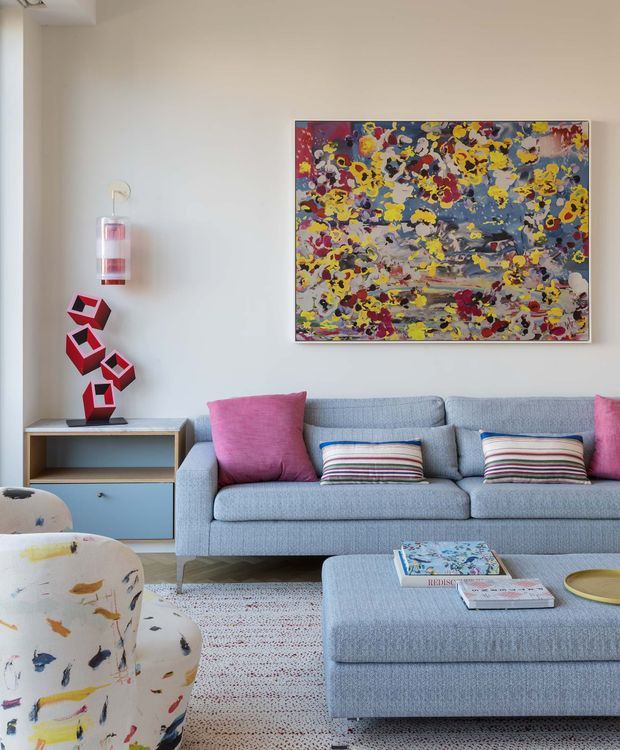 A bright, contemporary living room with a large light-blue sectional sofa and matching ottoman, colorful pink and striped cushions, and a patterned accent armchair. A vivid abstract artwork hangs above built-in blue and timber-toned cabinetry, accompanied by a sculptural red object. The room has a light neutral palette with playful, multicolored accents and abundant natural light.
