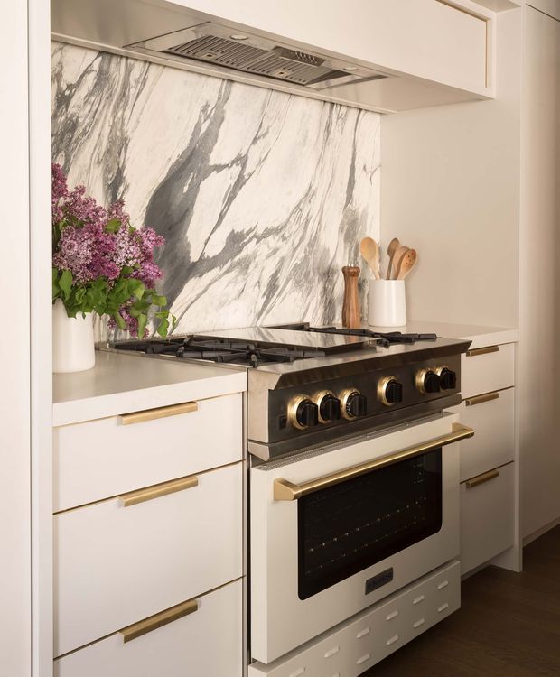 Close-up view of a refined kitchen cooking zone featuring a large freestanding range cooker beneath a stainless-steel rangehood. White cabinetry with slim brass-gold pulls surrounds the appliance, while a dramatic white-and-grey veined stone slab forms the backsplash. Neutral benchtops, timber flooring, a utensil holder, and a vase of flowers complete the warm, polished composition.