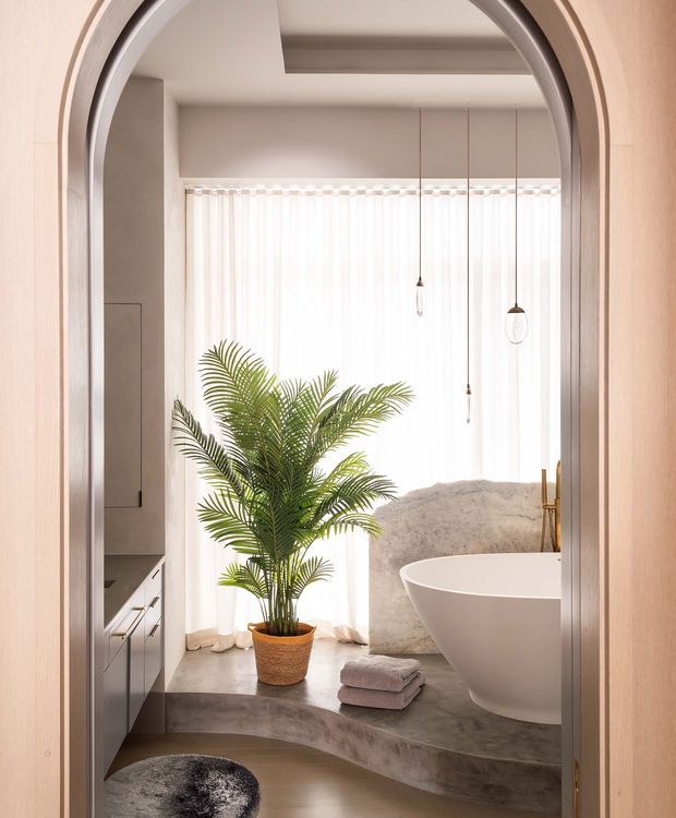 A softly lit bathroom viewed through a tall arched doorway. The room features a freestanding white oval bathtub on a raised curved concrete or stone platform, sheer floor-to-ceiling curtains, slim pendant lights, warm metallic tapware, grey cabinetry at left, and a potted palm. The palette is warm neutral with curved architectural forms and a refined contemporary feel.