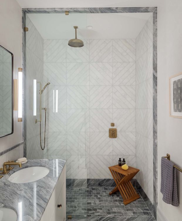 A refined bathroom centred on a frameless glass walk-in shower. The shower walls feature pale geometric patterned tile, complemented by dark grey stone-look floor tiles and a matching grey-veined stone vanity benchtop. Brushed brass shower fittings, tapware, and hardware add warmth, while a small timber stool and folded towel provide functional accents. A large mirror and narrow wall light are visible at the left.