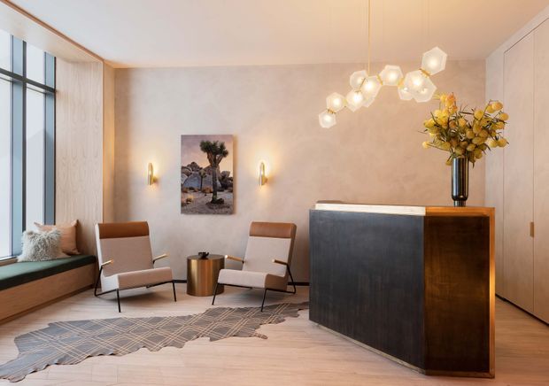 A refined residential entryway or living area with a built-in window seat, two mid-century-inspired lounge chairs, a sculptural pendant light, wall sconces, artwork, and a dark stone-and-timber console or bar cabinet. The room has warm timber flooring, pale textured walls, light built-in cabinetry, and a large black-framed window.
