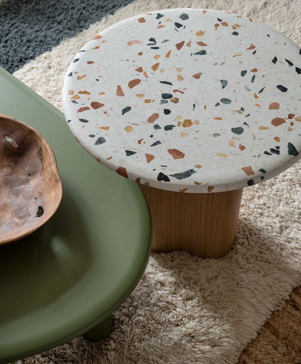 Close-up interior styling photograph of a rounded terrazzo-topped side table with multicoloured stone chips and a fluted or vertically textured timber pedestal base. It sits beside a sculptural olive-green upholstered chair and a natural wood bowl on a layered cream and grey shag rug. The composition highlights tactile materials, organic forms and earthy tones.