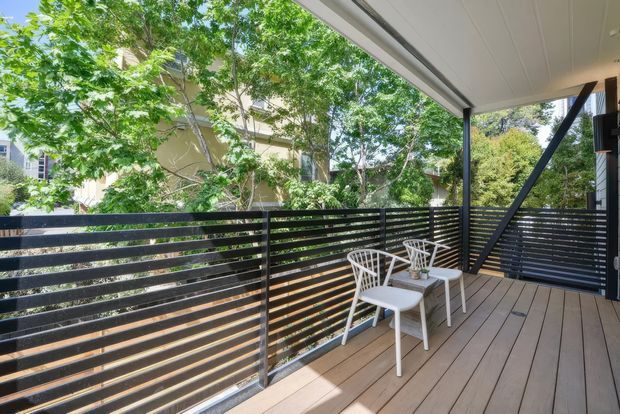 A bright covered outdoor deck or balcony with warm timber decking, black horizontal slatted privacy balustrades, and a black steel support frame. Two white outdoor chairs and a small matching table create a compact seating area, with leafy greenery and neighbouring homes visible beyond.