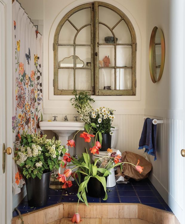 A compact, richly decorated bathroom viewed through an open doorway. The room features a white pedestal basin, dark blue square floor tiles, white vertical wall panelling, and an arched, weathered timber-framed mirrored cabinet or window above the basin. Floral-patterned fabric hangs at the left, while numerous potted flowering plants fill the floor and vanity area. An oval wall mirror, towels, and warm neutral walls complete the eclectic, vintage-inspired interior.
