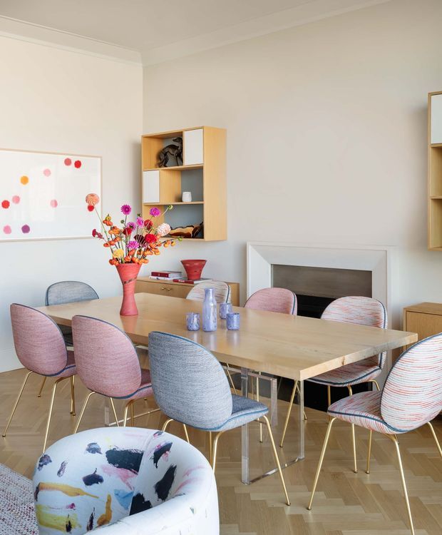Bright, stylish dining room with a large light-wood dining table surrounded by colourful upholstered chairs. The room has pale walls, parquet flooring, a simple white fireplace surround, built-in light-wood shelving, and colourful floral artwork and accessories, creating a playful contemporary interior.