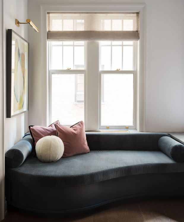 A compact, softly lit sitting area featuring a curved charcoal upholstered sofa beneath tall double windows. The windows have layered beige Roman shades and white painted trim, while blush cushions and a round cream cushion add restrained colour. A framed abstract artwork and brass wall-mounted reading light appear on the left wall, with dark timber flooring below.