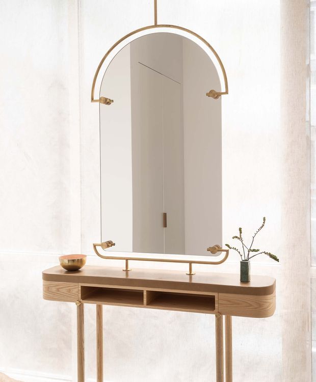 A refined interior vignette featuring a slim light-wood console table with rounded edges, open central shelving, and slender legs. Above it is a large arched mirror with a distinctive brass or bronze frame and matching visible mounting hardware. Sheer curtains provide a softly lit backdrop, while a small bowl and narrow vase with branches sit on the console. The overall composition is minimalist, warm, and elegant.