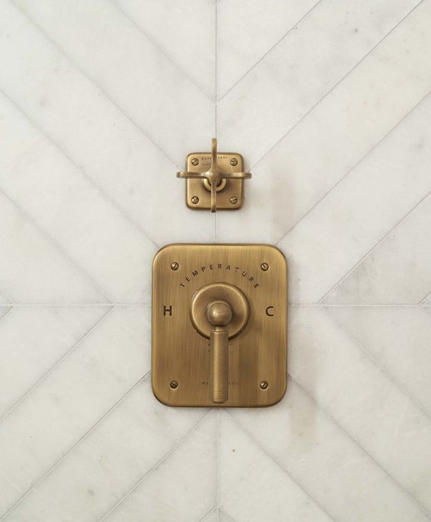 Close-up of a shower control and diverter mounted on a white marble-tiled wall. The fixtures have a warm brushed brass or bronze finish, with a vintage-inspired rectangular control plate marked with hot and cold indicators and a matching upper diverter.