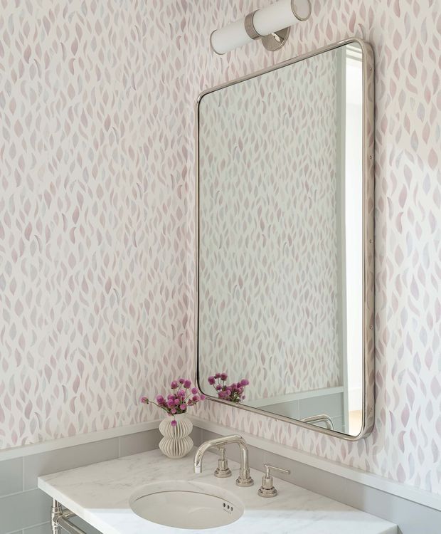 A refined bathroom vanity vignette with a large rounded-rectangle mirror, pale marble countertop and integrated oval sink, polished metal tapware, and a small ribbed ceramic vase with purple flowers. The walls are covered in a soft pink-and-white leaf-patterned finish, complemented by pale grey wainscoting and a cylindrical wall light.