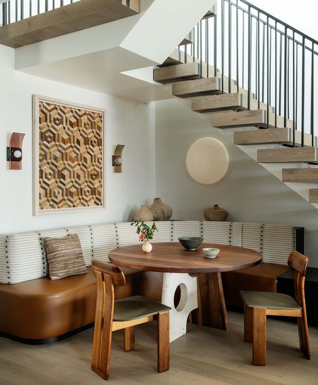 A refined dining nook beneath an open staircase, featuring a built-in curved banquette with striped upholstery, a round dark timber dining table, timber dining chairs, and sculptural ceramic decor. The room has warm timber flooring, white walls, framed geometric artwork, wall sconces, and a contemporary staircase with floating timber treads and slender dark metal balustrades.