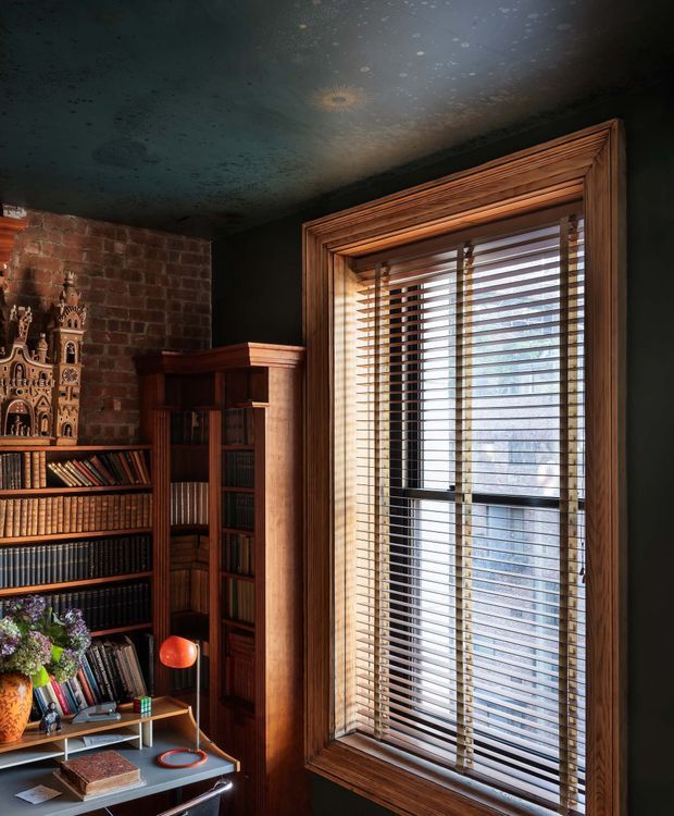 A richly detailed, moody home library or study corner with dark painted walls and ceiling, exposed red brick, extensive dark timber bookcases, and a large timber-framed window fitted with horizontal blinds. Books, a decorative clock, flowers, and a small desk with a bright orange lamp create a warm, eclectic atmosphere.