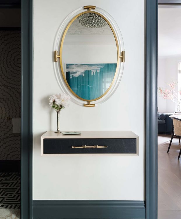 A refined interior entry vignette featuring a wall-mounted floating console with a pale stone or light solid-surface top, dark drawer front, and brass handle. Above it is a large oval mirror with a white and brass frame, reflecting a textured ceiling light and blue-toned artwork. Dark blue-grey door trim and wall panelling frame the composition, with glimpses of adjacent rooms at either side.