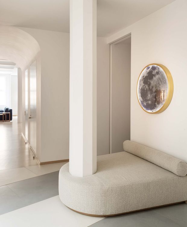 Bright, refined interior view of a contemporary hallway and seating nook. A curved cream upholstered built-in or fitted chaise wraps around a substantial white structural column, with a slim timber-toned base detail. The space has pale smooth plaster walls and ceilings, large light-grey floor tiles, white doors and trims, and a rounded arched ceiling transition visible along the corridor. An oval gold-framed artwork hangs above the seating, while a brighter room with additional seating is visible down the hall.