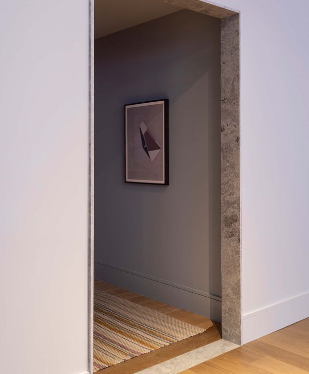 A moody interior view through a wide wall opening into a narrow hallway or entry passage. The opening is framed with a rough, mottled stone or concrete-like surround, while the surrounding walls are painted soft white and charcoal grey. A framed abstract artwork hangs on the rear wall above a low skirting board. Timber flooring continues into the passage, with a woven striped runner partly visible, creating a refined, contemporary transitional feel.