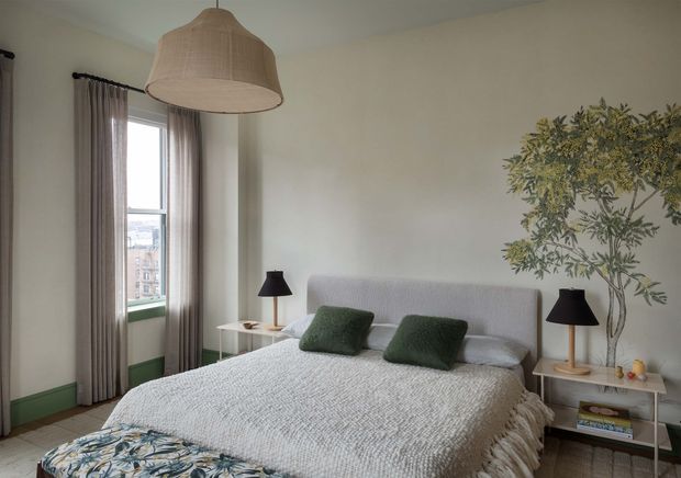 A calm, softly styled bedroom with a large upholstered bed, textured pale coverlet, dark green cushions, and a floral upholstered bench at the foot. Tall windows with sheer taupe curtains sit beside the bed, while matching black table lamps, a woven pendant light, green-painted skirting, and a leafy indoor tree add character.