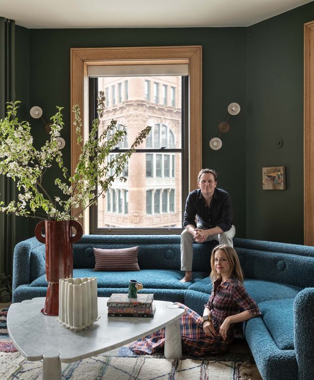 A richly styled living room with deep green walls, warm timber window and door trims, and a large city-facing window. A sculptural blue sectional sofa wraps around a pale stone coffee table, accompanied by a patterned rug, decorative vase, books, plants, and sculptural wall lights. Two people are seated in the room. The space has a sophisticated, moody yet inviting character with a blend of contemporary and retro-inspired furnishings.