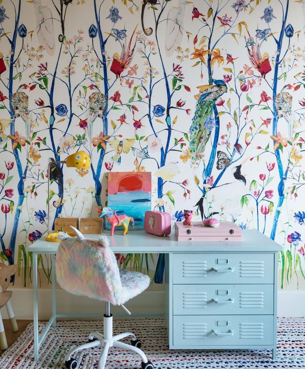 A playful children's study or bedroom corner with a pale blue metal desk featuring drawers, a pastel faux-fur swivel chair, colourful toys and artwork, and an expressive full-height botanical and bird mural wallpaper. The room has a bright, whimsical, highly colourful interior with a patterned rug and light neutral background.