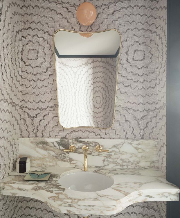 A compact, highly decorative bathroom vanity featuring a sculptural white inset basin set into a richly veined marble countertop and backsplash. The wall is covered in a monochromatic scalloped floral patterned finish, complemented by a rounded rectangular mirror with a slim brass frame, brass wall-mounted tapware, and a soft peach globe wall light. The overall composition is symmetrical, refined, and glamourous.