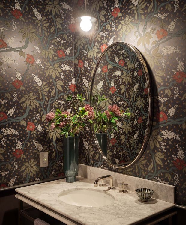 A richly styled powder-room vanity with dark botanical floral wallpaper, a large oval mirror, and a marble countertop with an integrated white basin. A dark wood vanity, polished metal tapware, wall sconce, and floral arrangement create a dramatic, vintage-inspired interior.
