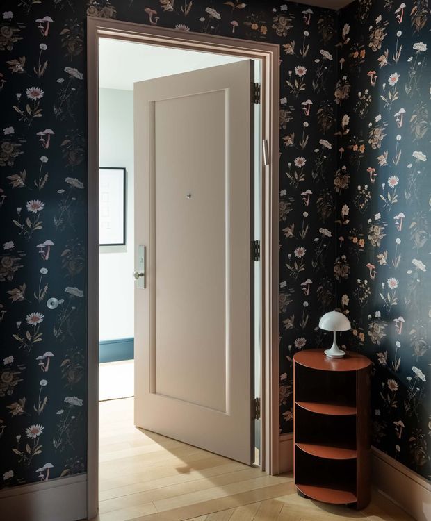A compact, richly decorated entryway or hall with dark botanical floral wallpaper, timber skirting, and a warm wood-toned curved shelving console topped with a white dome table lamp. A paneled cream interior door stands open to a brighter adjoining room with pale timber flooring and blue lower wall detailing. The space has a strongly patterned, vintage-inspired yet polished character, with sunlight entering from the adjoining room.