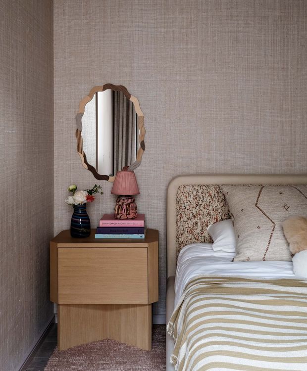 A close-up view of a warmly styled bedroom corner featuring a timber bedside table, decorative mirror, lamp, books, flowers, and the edge of a neatly made bed. The room has textured taupe wall covering, a padded upholstered headboard, layered neutral bedding, and soft earthy accents.