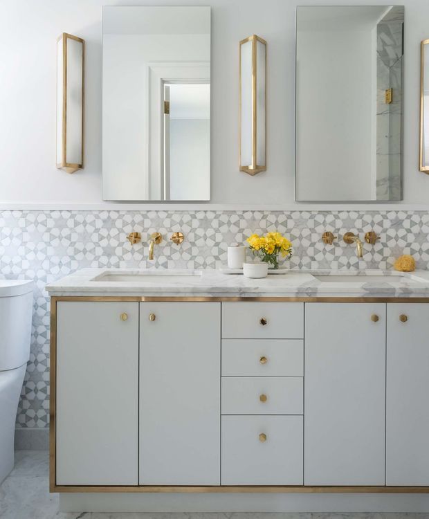 A bright, refined bathroom vanity with a wide grey painted cabinet, marble benchtop and integrated basins. The wall is finished with geometric patterned mosaic tile, complemented by brass wall-mounted tapware, brass-framed vertical sconces, mirrored cabinets, and gold-toned hardware. A toilet is partially visible at left.