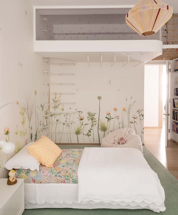 A bright, playful bedroom with a low white bed dressed in scalloped white bedding, a colourful floral-patterned coverlet, and a gingham accent cushion. The walls feature delicate botanical illustrations, with open white shelving and built-in storage above the bed. A sculptural woven pendant light hangs near the ceiling, while a plush rounded chair and bookshelves are visible to the side. The room has a soft pastel palette and a whimsical, child-friendly atmosphere.