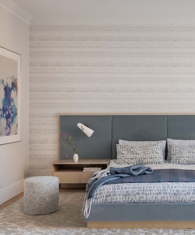 A refined contemporary bedroom featuring a low upholstered bed with a large charcoal-blue padded headboard, integrated timber bedside joinery, and a wall-mounted reading lamp. The room has pale textured horizontal wallcovering, patterned blue-grey bedding, a speckled upholstered stool, light timber flooring, and abstract blue artwork. The palette is soft and muted with layered grey-blue and neutral tones.