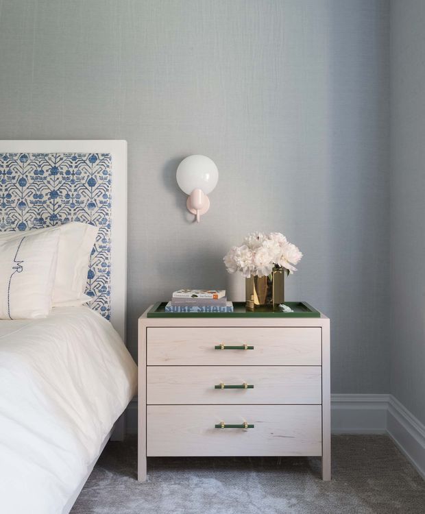 A softly styled bedroom corner featuring a white upholstered bed with a blue-and-white patterned headboard, pale blue-grey textured walls, white trim, grey carpet, and a light timber three-drawer bedside chest with green-and-brass pulls. A globe wall sconce and floral arrangement add decorative accents.