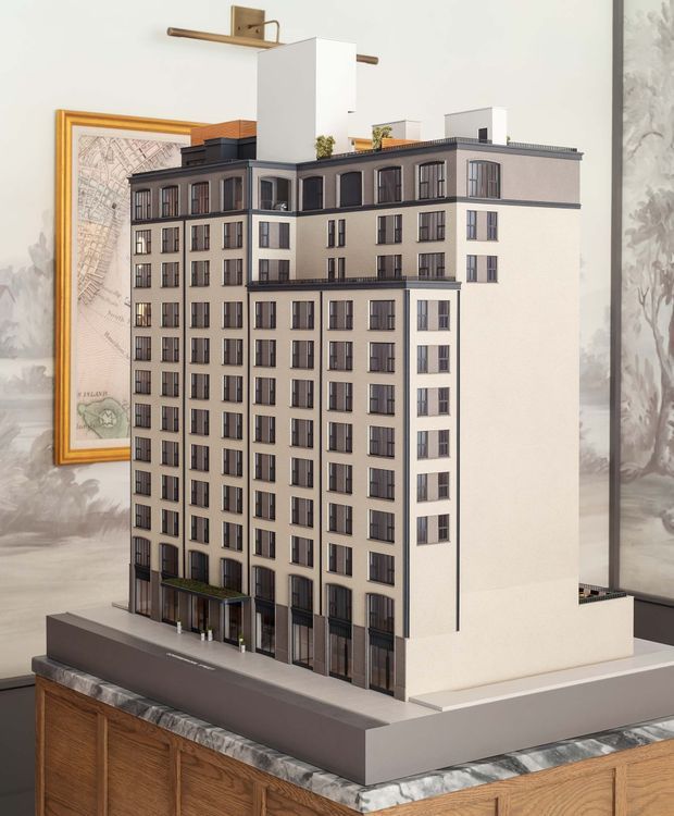 A detailed architectural scale model of a multi-storey urban apartment building displayed on a large rectangular base. The model has a cream and grey facade, repetitive dark-framed windows, projecting cornices and balconies, rooftop structures and planting, and a ground-level entrance canopy. It is photographed indoors on a timber cabinet, with framed artwork and patterned wall decor in the background.