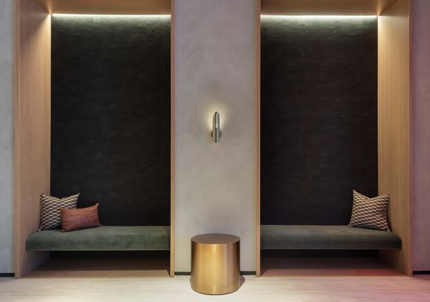 A symmetrical, built-in seating alcove with two upholstered bench seats set into warm timber niches. Dark textured wall panels create a dramatic backdrop, while concealed linear lighting highlights the upper edges. A central pale textured partition features a small wall light, with a cylindrical metallic side table below. The design is refined, moody, and highly tailored.