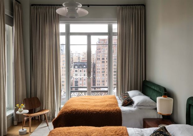 A refined bedroom with a large city-facing window and balcony doors framed by full-height taupe curtains. The room contains a bed with a deep green upholstered headboard, white bedding, rust-orange textured throws, a sculptural white pendant light, a small timber bedside table, and a mid-century-style wooden chair. Urban apartment buildings are visible beyond the glazing.