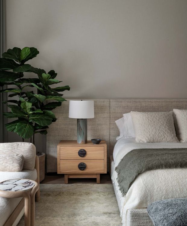 A softly styled bedroom corner with a bed on the right, a wooden two-drawer bedside table and lamp at center, and a large leafy indoor plant beside a partial upholstered armchair on the left. The room uses layered neutral textiles, a textured upholstered headboard, a muted rug, and warm natural wood accents, creating a calm contemporary look.