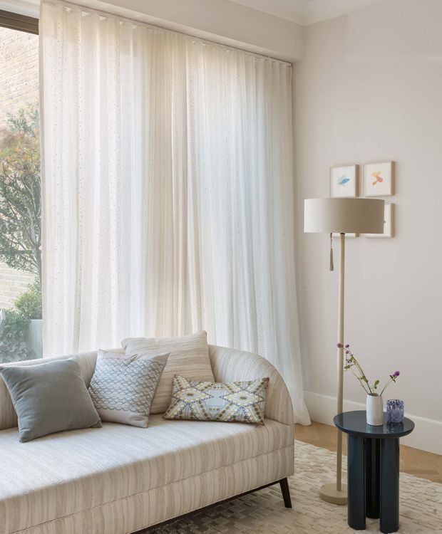 A softly lit living room corner with a curved upholstered sofa, layered patterned cushions, sheer full-height curtains over a large window, a slim brass floor lamp, small round black side table, framed artwork, and a light textured rug. The palette is predominantly warm white, cream, beige, and pale grey with subtle decorative accents, creating a calm contemporary interior.
