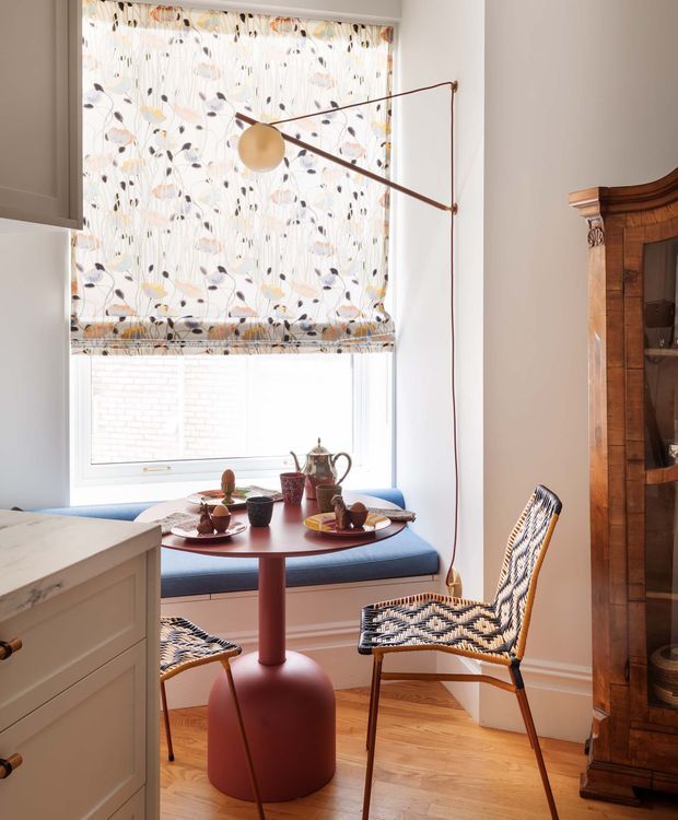A bright, compact dining nook beside a large window, furnished with a round reddish table, blue built-in bench seating, and woven patterned chairs. A floral Roman blind, sculptural wall-mounted reading lamp, timber parquet floor, white trim, painted cabinetry, and a large traditional timber display cabinet create a colourful, eclectic interior.