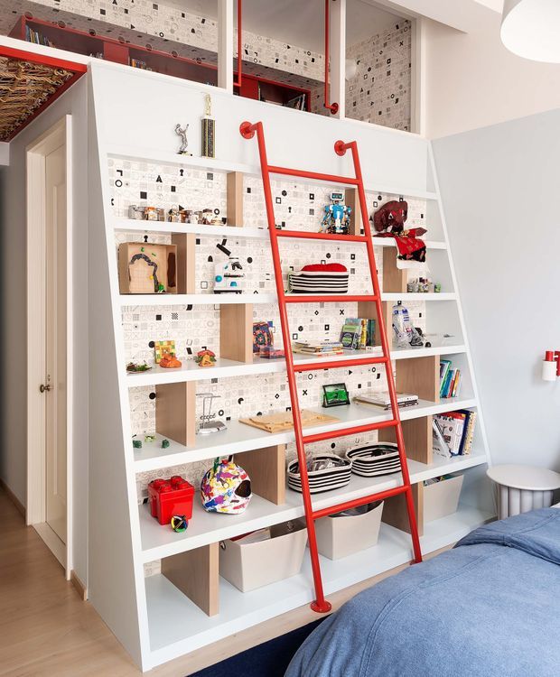 Bright children's bedroom featuring a large custom white built-in shelving and display unit spanning the wall, with open cubbies, pale timber backing sections, and playful patterned wallpaper. A vivid red fixed ladder provides access to upper storage, while colourful toys, books, baskets, and objects fill the shelves. Part of a bed with blue bedding is visible in the foreground, and a small round stool sits to the right. The room has light timber flooring and a clean, playful contemporary fit-out.